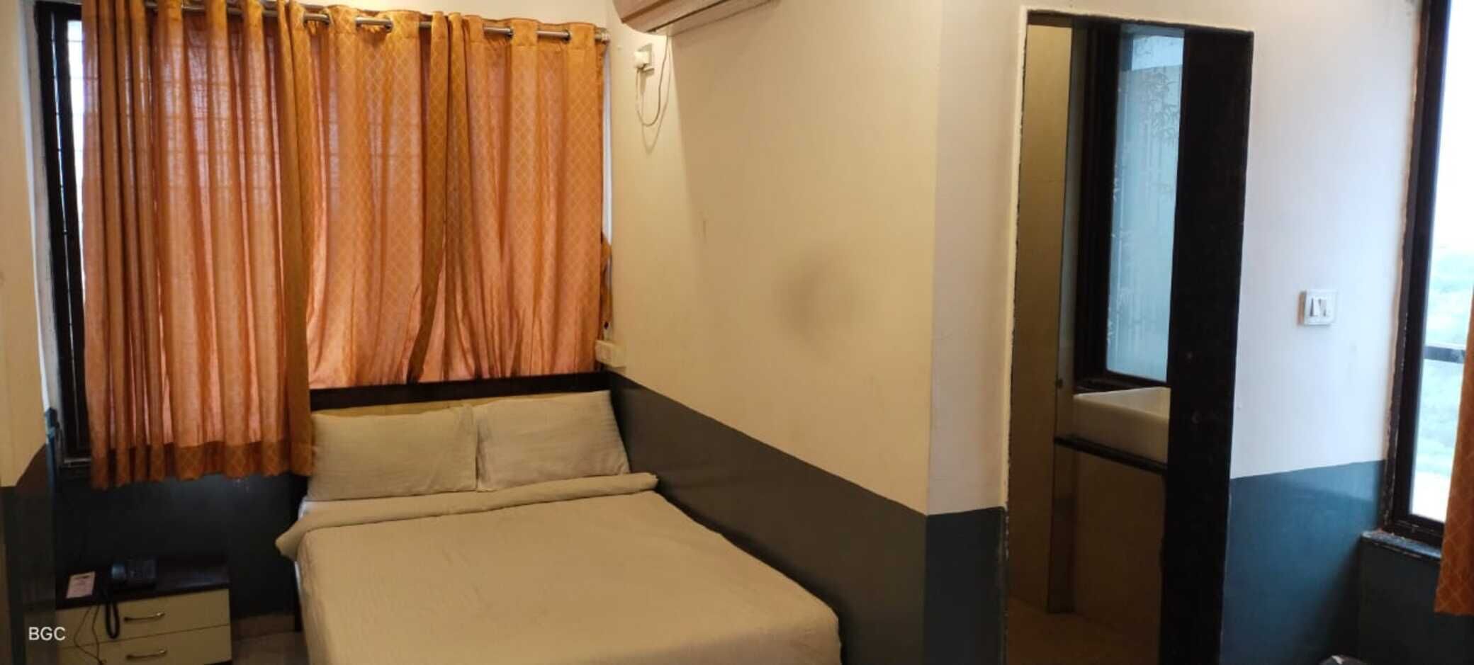 Pequeña habitación individual con cama, mesita de noche, ventana con cortinas y baño a la vista a través de una puerta abierta.