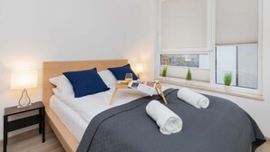 Una cama bien hecha con una colcha gris, dos almohadas azules y un cabecero de madera. Hay una mesita de noche con una lámpara, una bandeja con un vaso de jugo de naranja y una revista. Se ve una ventana con persianas y una pequeña planta en el alféizar.