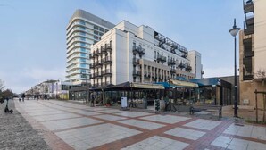 A modern building with multiple floors, a covered entrance, and a paved plaza with benches. - Sunny Apartment with Parking | Near the Beach | Air Conditioning (Świnoujście)