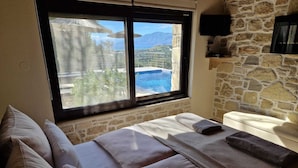 Une pièce avec un mur en pierre, un lit avec des oreillers, une table de massage avec des serviettes, et une fenêtre donnant sur une piscine et des montagnes.