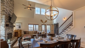 A spacious living area with a fireplace, a dining table set for four, and a staircase leading to another level. - Woof Lodge at SweetGrass - pet friendly, outdoor fireplace, walk to lake (Blowing Rock)