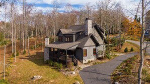 A two-story house with a stone chimney, a porch, and a driveway leading to the entrance. - Woof Lodge at SweetGrass - pet friendly, outdoor fireplace, walk to lake (Blowing Rock)