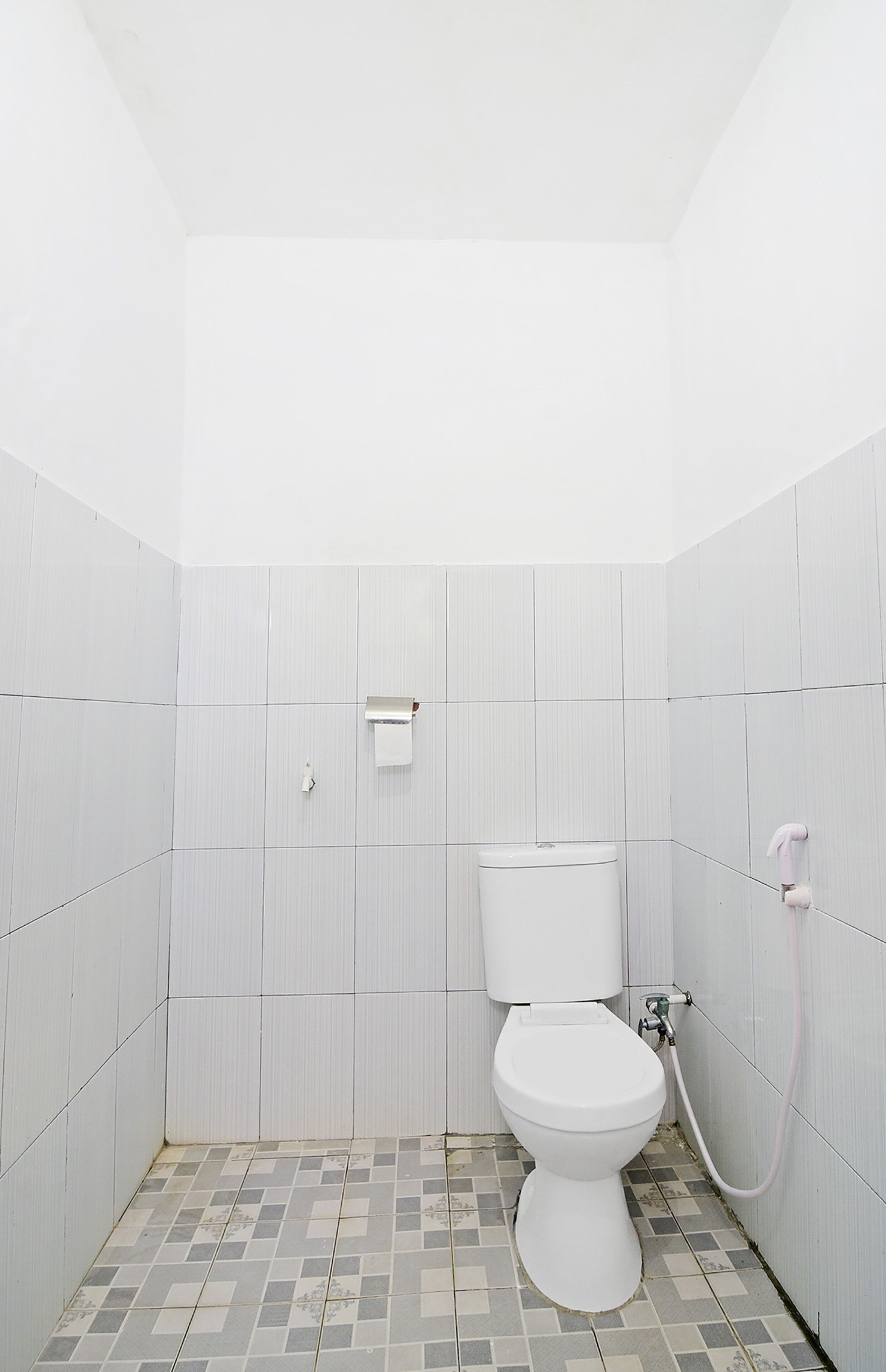 Un baño pequeño con azulejos blancos, un inodoro, un dispensador de toallas de papel y una tubería visible.