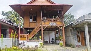 A two-story wooden house with a balcony, a sign reading "Juniper Home Sweet," and various potted plants around the property.
