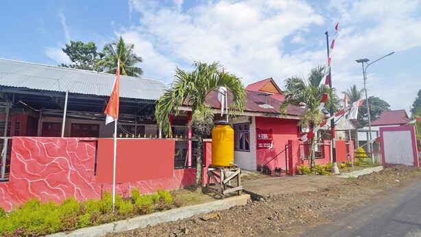 A building with a red exterior and a corrugated metal roof, surrounded by greenery and palm trees. - Hotel O Pulisan Ekowisata Homestay Near Wisata Pantai Desa Pulisan (Pulisan Beach Tour) (Manado)