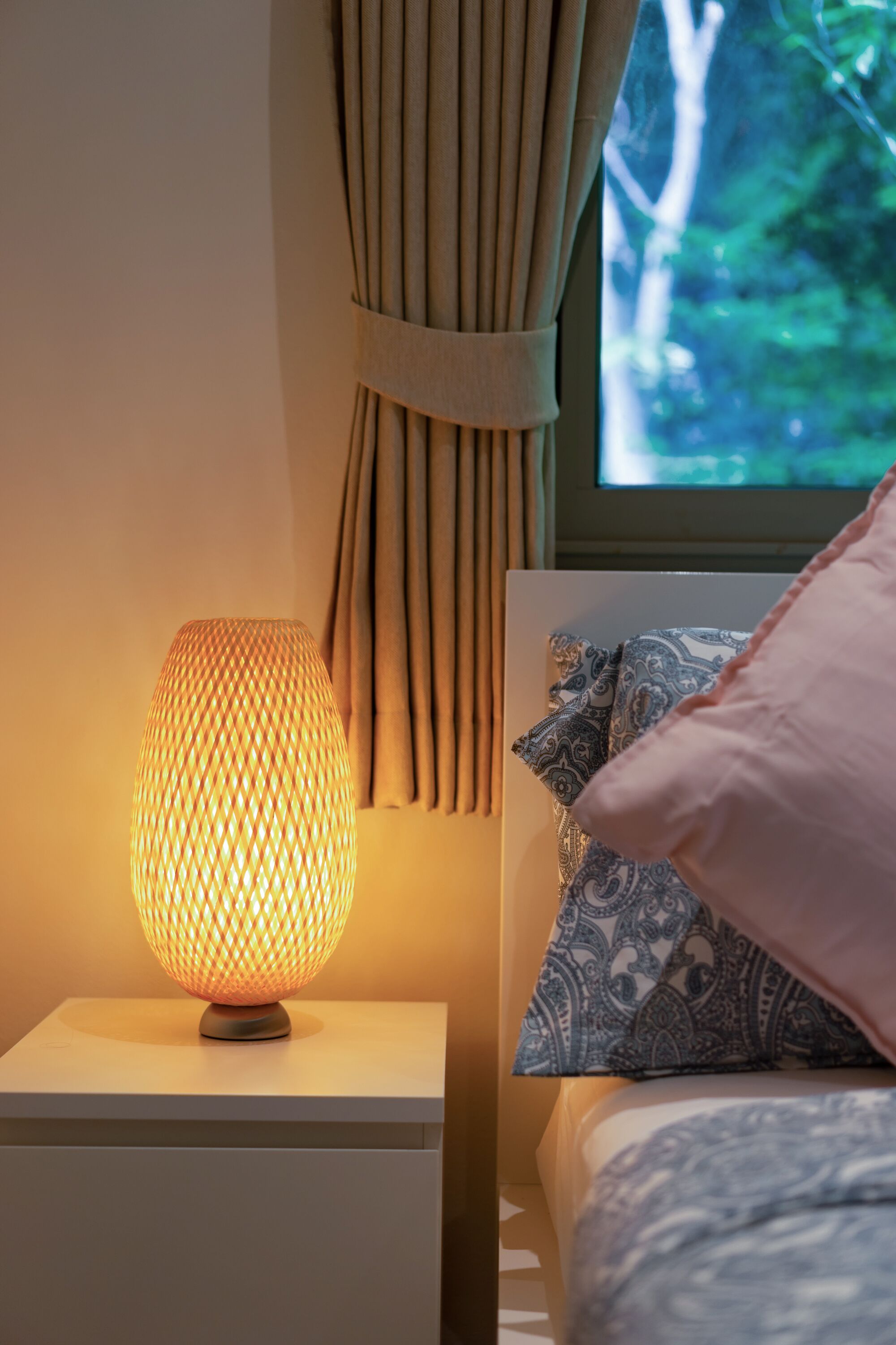 A bedside lamp with a textured shade, a patterned pillow, and a view of greenery through a window.