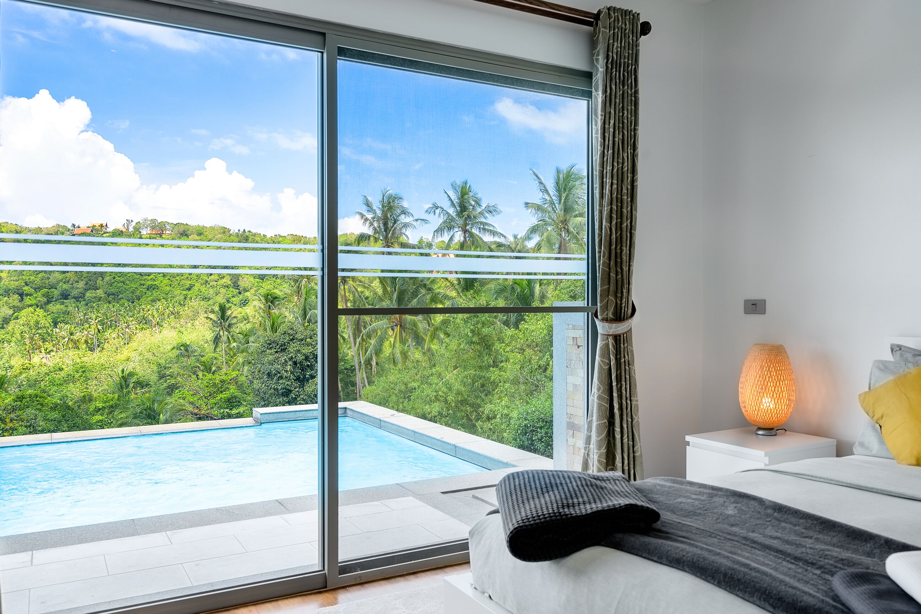 A bedroom with a large window overlooking a swimming pool and lush greenery.