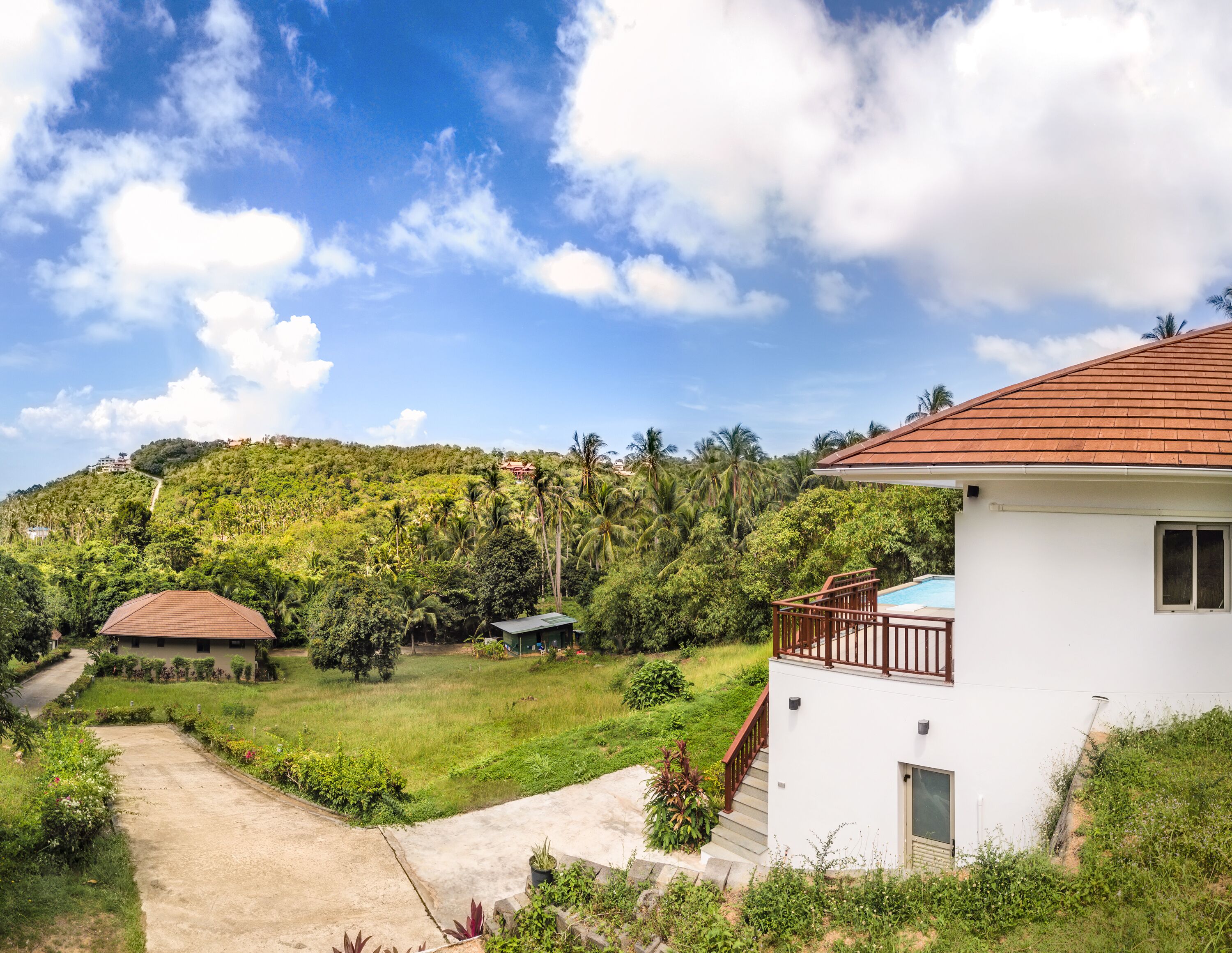 A white building with a red roof, a small pool, and a garden area with a pathway leading to a hilltop house.