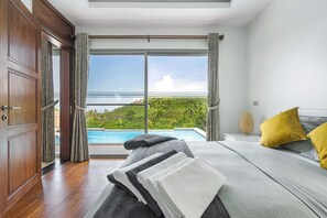 Une chambre d’hôtel avec une grande fenêtre, un lit avec une literie grise, un oreiller jaune et une vue sur une piscine et de la verdure.
