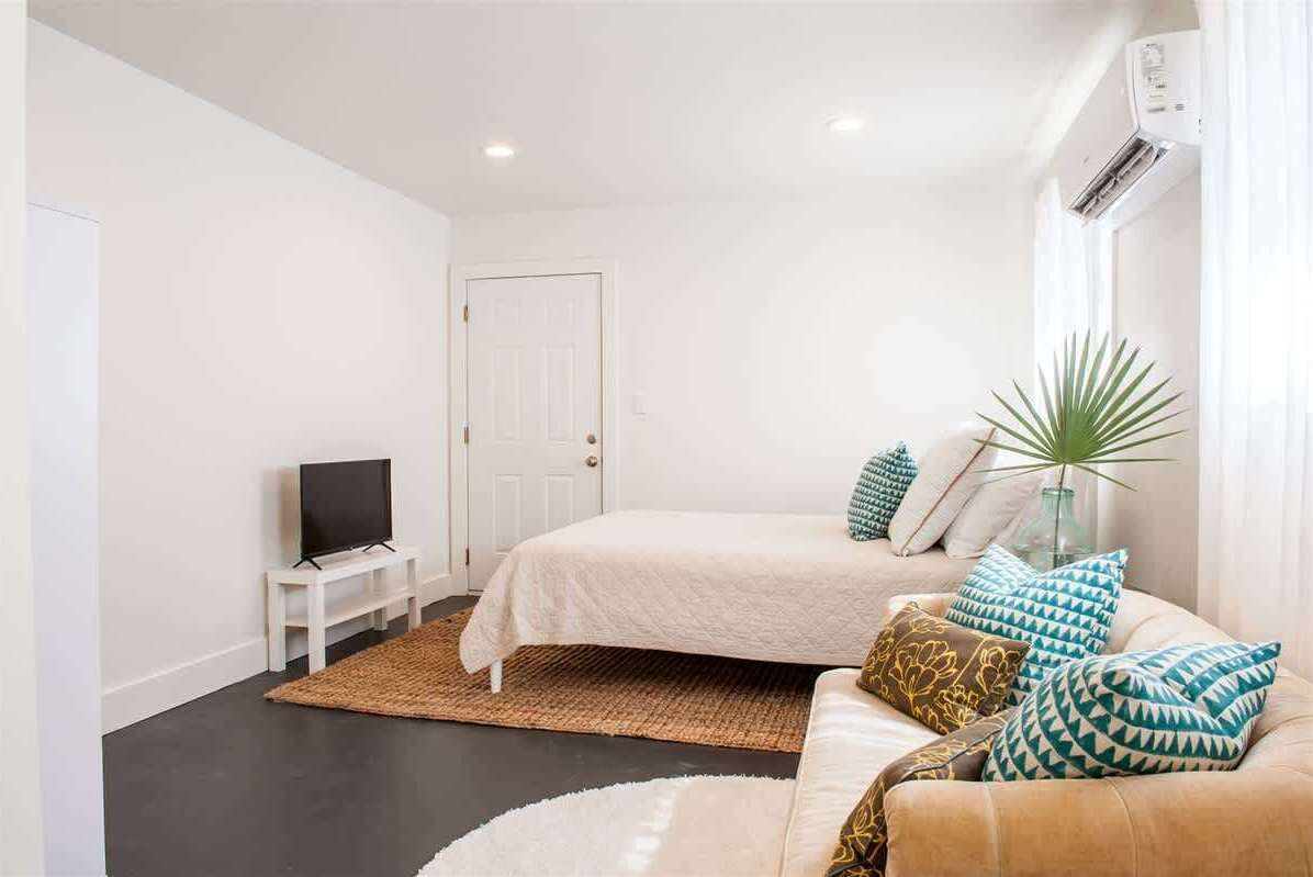 A bedroom with a bed, a TV on a stand, a sofa, a plant, and an air conditioning unit.