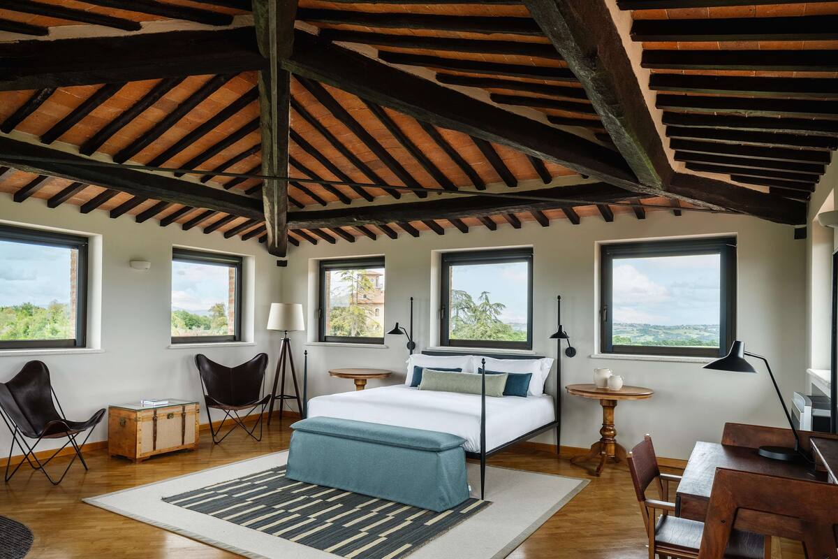 Ein Schlafzimmer mit einem großen Bett, Holzbalken, mehreren Fenstern, einem Schreibtisch und einem Stuhl.