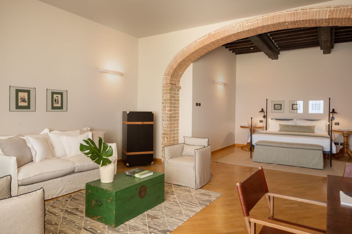 Ein modernes Wohnzimmer mit einer Couch, einer grünen Kommode, einem Couchtisch mit Pflanze und einem Ziegelbogen, der in ein Schlafzimmer mit Bett und Nachttisch führt.