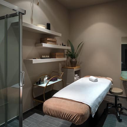 A modern bathroom with a glass shower enclosure, a bed with a white towel, shelves with toiletries, and a sink with a mirror.