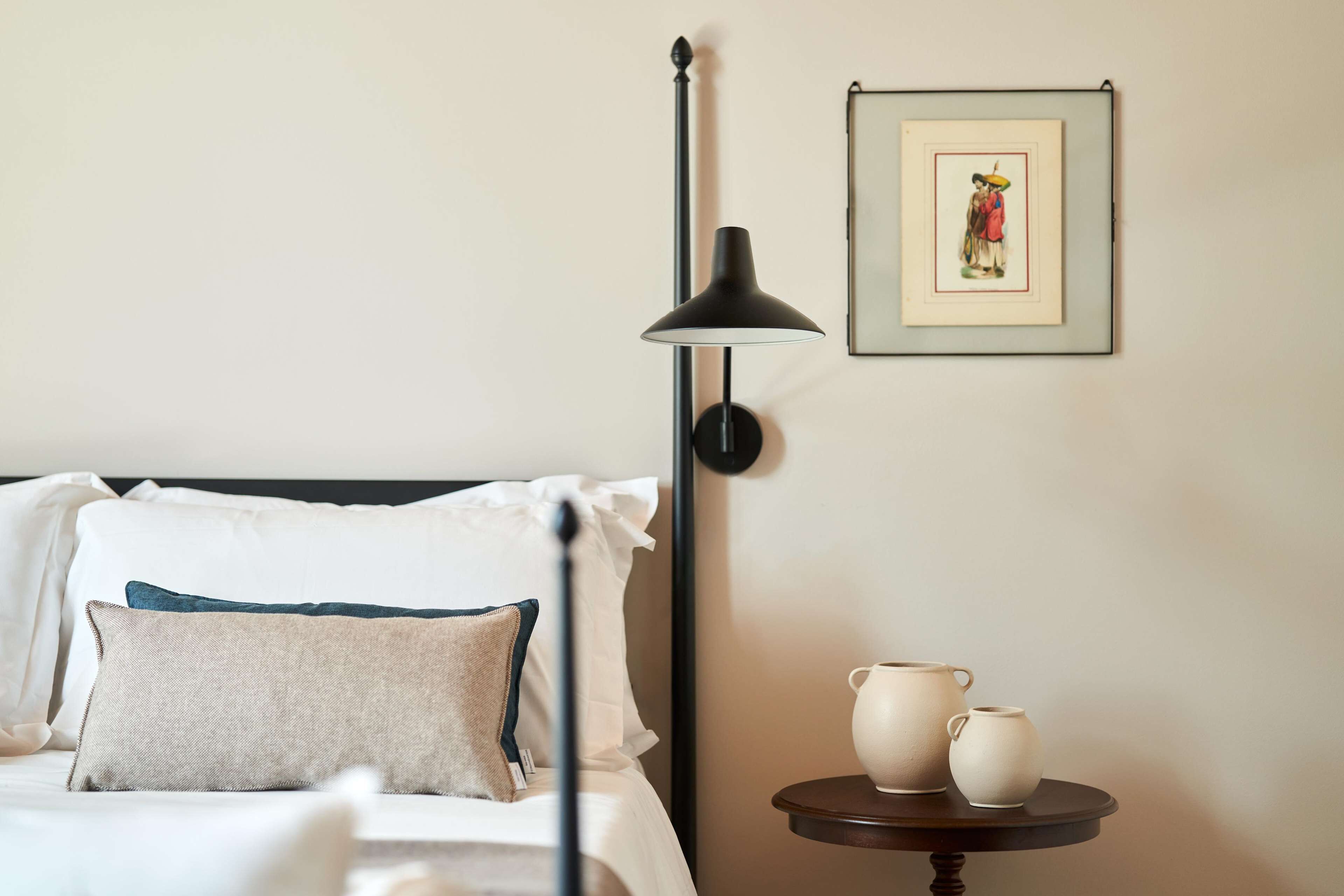 A neatly made bed with pillows, a floor lamp, a framed picture on the wall, and a small table with two ceramic cups.