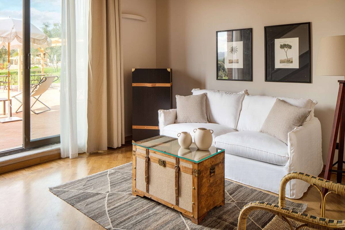 Ein Wohnzimmer mit einem weißen Sofa, einem Glastisch mit zwei weißen Karaffen, einem Korbstuhl und einem Weidenkorb.