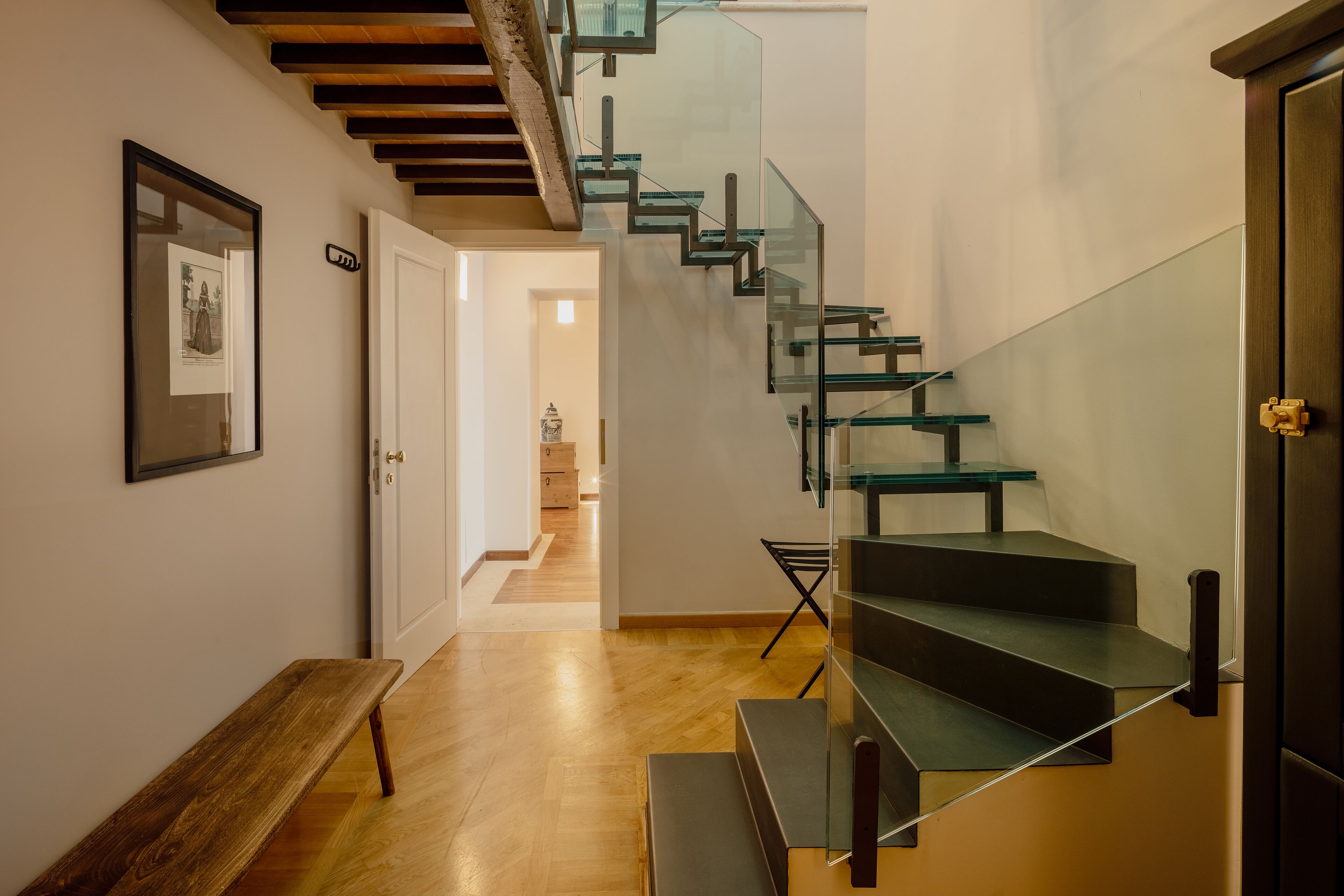 A modern staircase with glass railings and wooden steps, leading to a room with a bench and a framed picture on the wall.