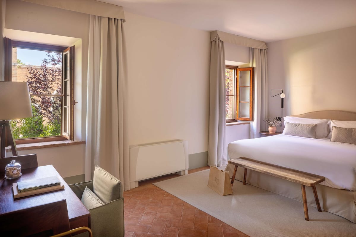 Ein Hotelzimmer mit einem Bett, einem Schreibtisch mit Stuhl, einem Fenster mit Vorhängen, einem Heizkörper und Blick auf Grünflächen.