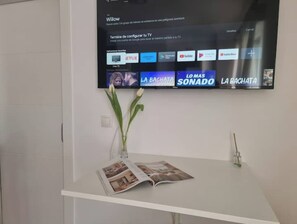 A reception desk with a magazine, a vase with flowers, and a TV displaying streaming services. - 600m de la Playa, Terraza Soleada (Salobreña)