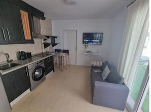 A compact kitchen with a washing machine, a dining area with a small table and chairs, a sofa, and a TV mounted on the wall. - 600m de la Playa, Terraza Soleada (Salobreña)