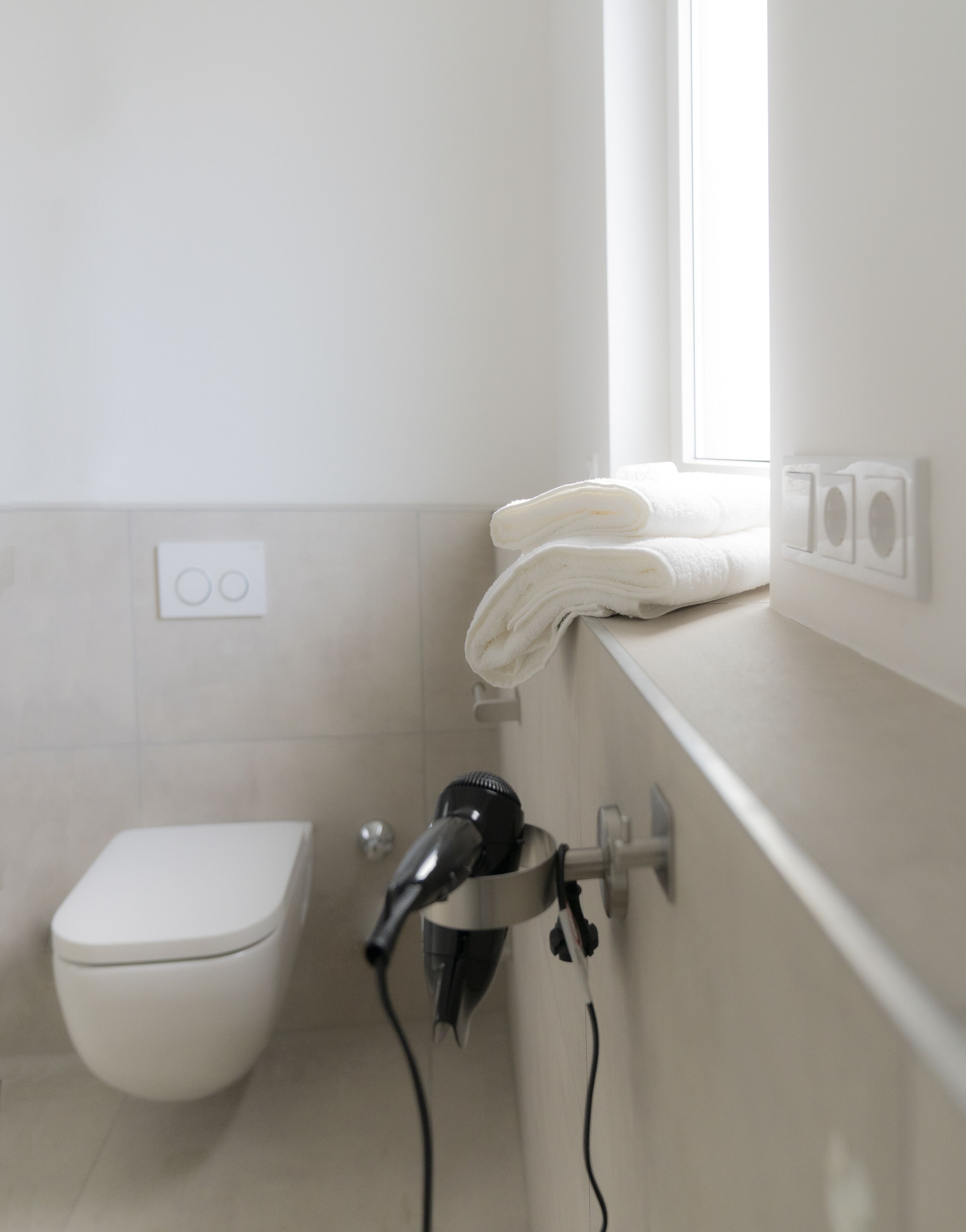 Een badkamer met een wit toilet, een haardroger op een wandhouder, en een raam voor natuurlijk licht.