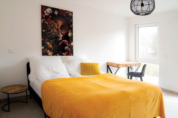 Une chambre à coucher comprenant un lit, une petite table, une chaise, une fenêtre et une œuvre d’art accrochée au mur.