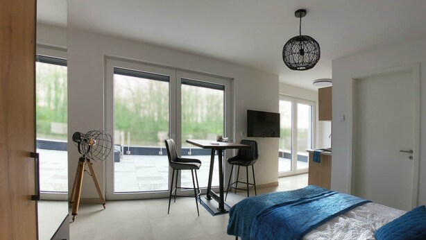 A modern bedroom with a bed, a desk, two chairs, a TV, and large windows. - Penthouse apartment no. 10 on the 2nd floor on the right (garden view, large roof terrace) (Dormagen)