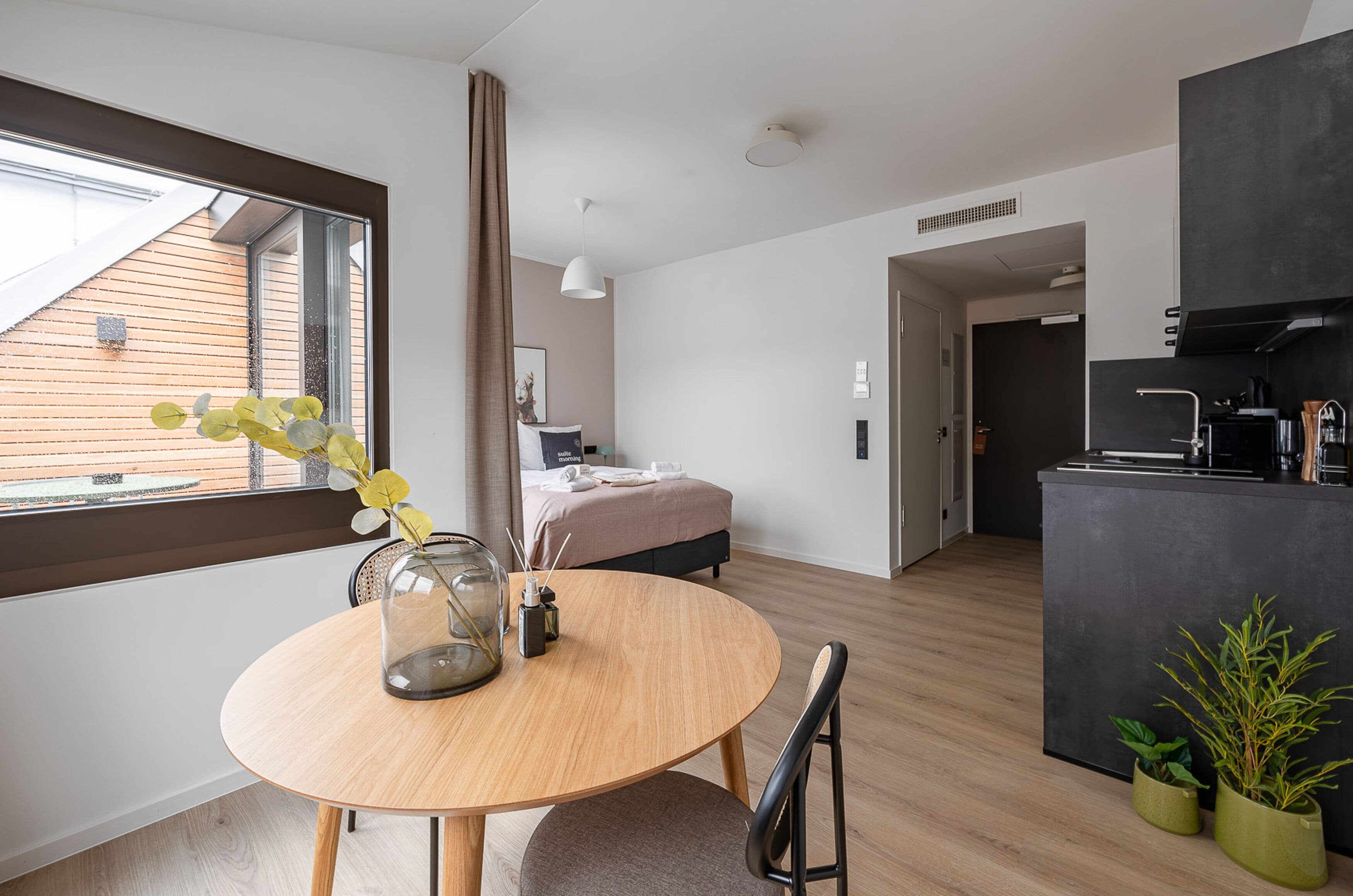 A modern hotel room with a round wooden dining table, a bed with a beige headboard, a kitchen area, and a view of the outside through a large window.