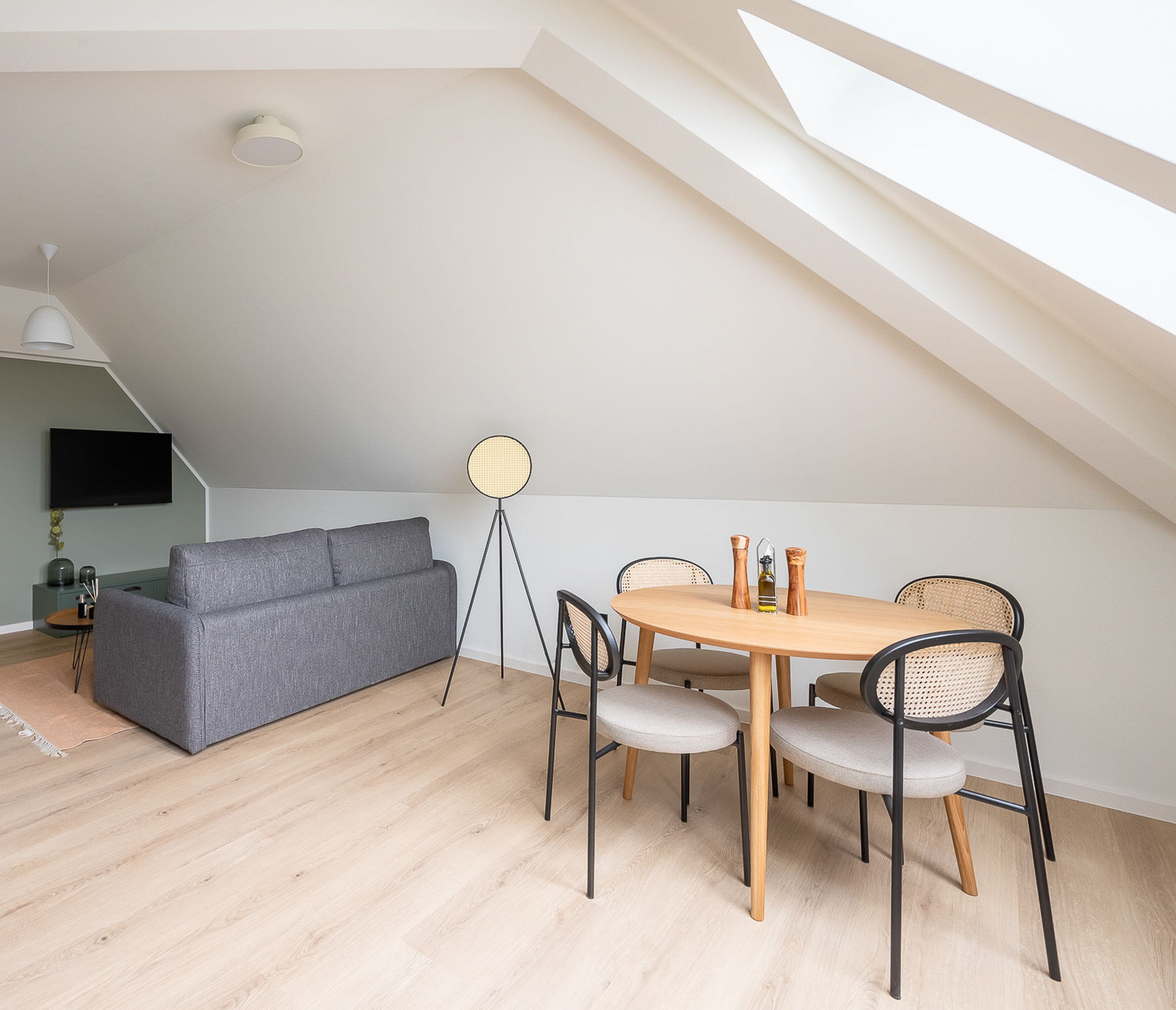A modern loft with a dining area featuring a round wooden table and four chairs, a grey sofa, and a floor lamp.