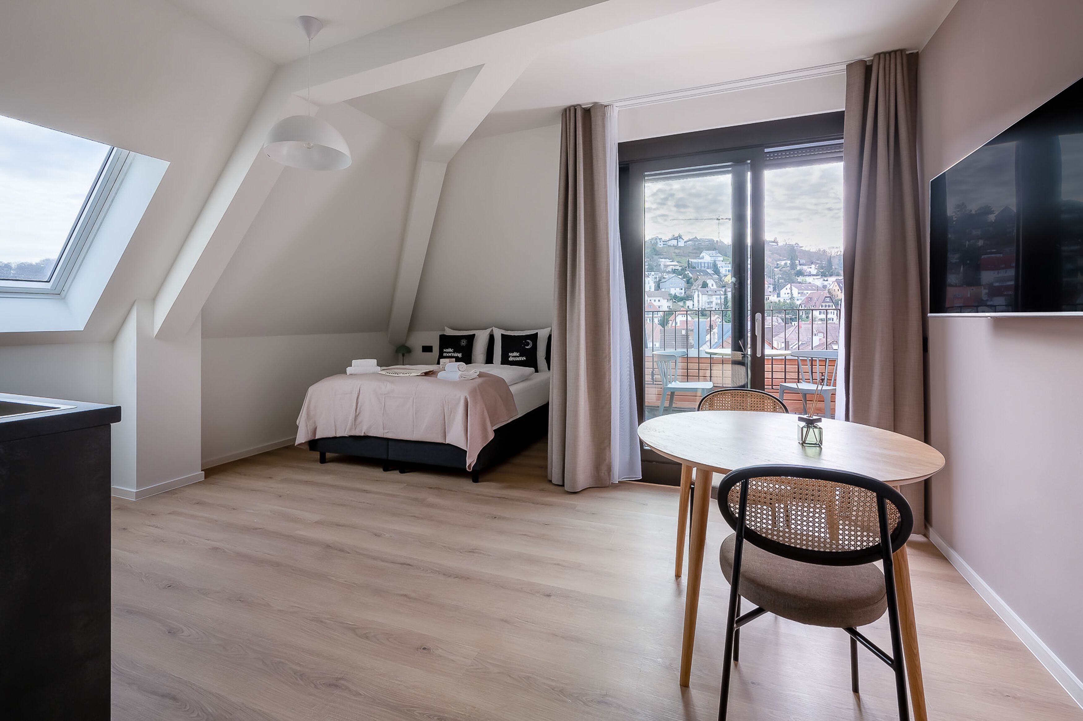 A modern bedroom with a skylight, a bed, a dining table, and a view of the city through large windows.