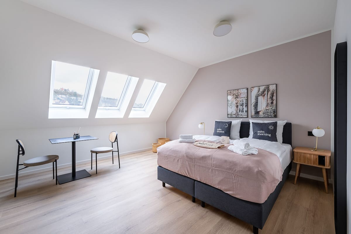 A modern bedroom with a bed, two chairs, a small table, and a desk. There are skylights and a view of the city.
