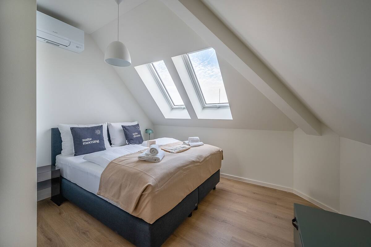 A modern bedroom with a skylight, a bed with a beige blanket, and a "suite morning" pillow.