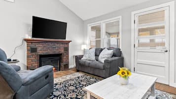 A living room with a fireplace, a grey sofa, a coffee table with a vase of sunflowers, and a patterned rug.