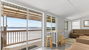Ein modernes Zimmer mit einer großen SchiebeglasTür, die auf einen Balkon mit Blick auf ein Gewässer und die Ferne führt. Es gibt einen Holztisch mit zwei Hocker und ein beiges Sofa.