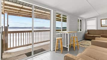 A modern room with a large sliding glass door leading to a balcony with a view of a body of water and distant land. There is a wooden table with two stools and a beige sofa.