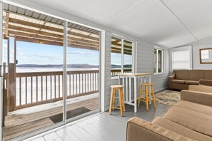 A modern room with a large sliding glass door leading to a balcony with a view of a body of water and distant land. There is a wooden table with two stools and a beige sofa. - 2BR Camp w/ Amazing Beach Views (Colchester)