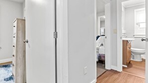 Ein Zimmer mit Holzschrank, einem Bett mit weißer, gemusterter Bettdecke und einem Badezimmer mit weißem Waschbecken und Spiegel.