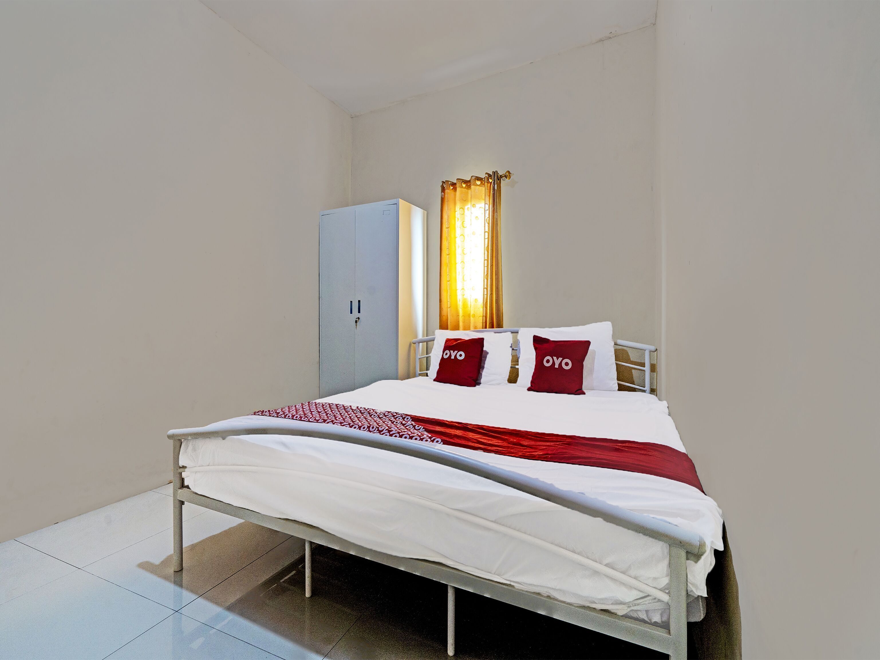 A single bed with white bedding and red pillows, a grey wardrobe, and a window with curtains.