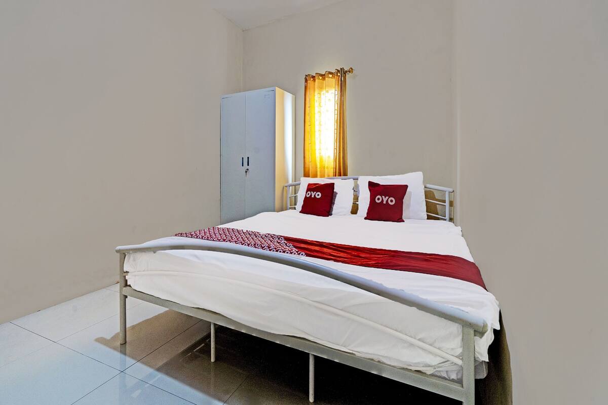 Una cama individual con ropa de cama blanca y almohadas rojas, un armario gris y una ventana con cortinas.
