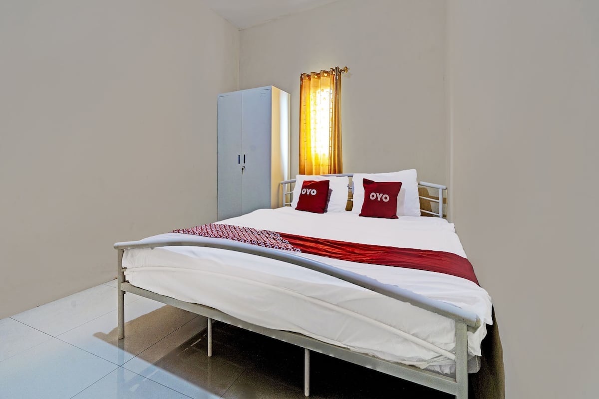 Una cama individual con ropa de cama blanca y almohadas rojas, un armario gris y una ventana con cortinas.