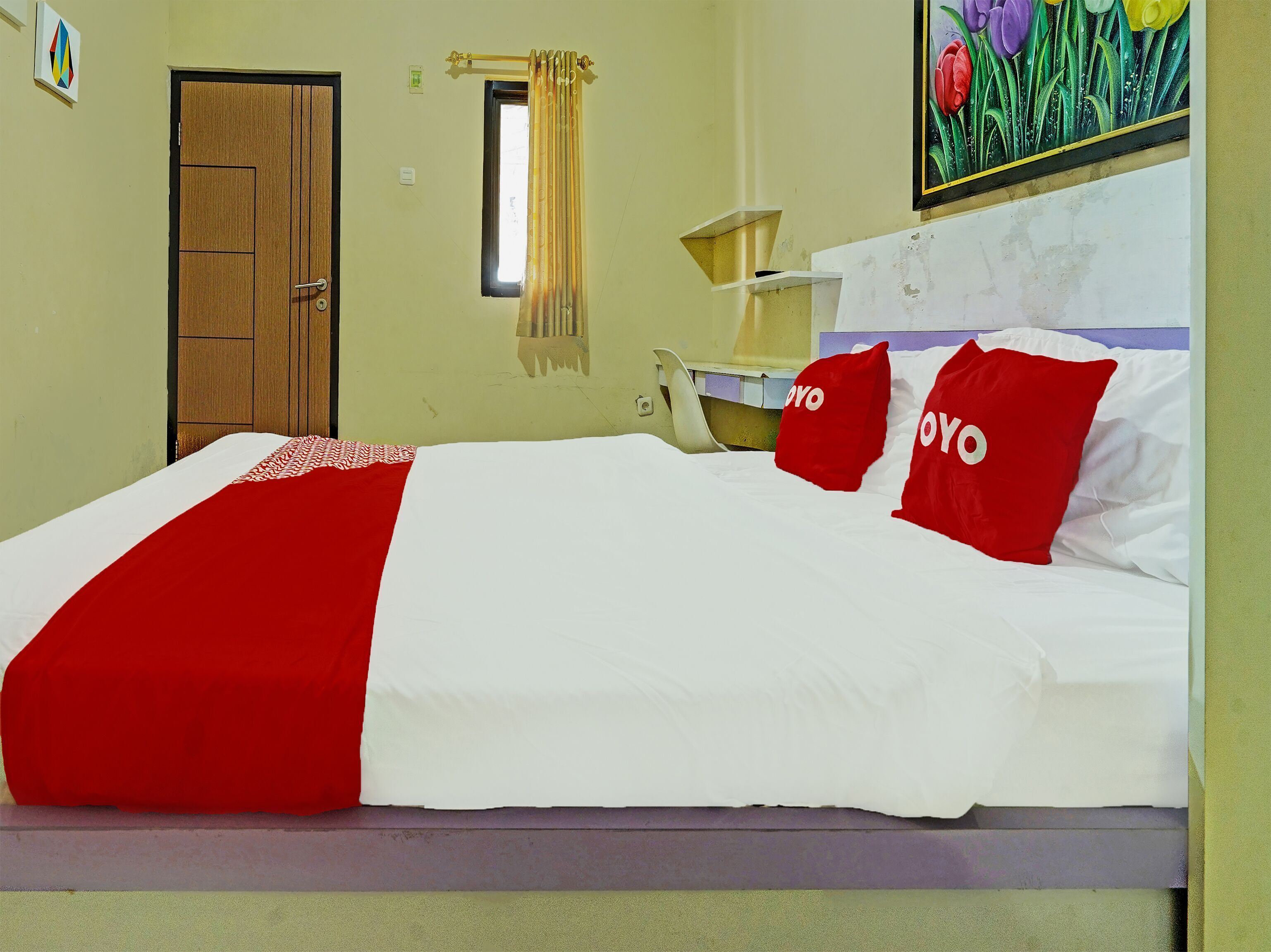 A bed with a red and white bedspread, two red pillows with 'YOYO' text, a window with a curtain, a door, and a painting of flowers on the wall.