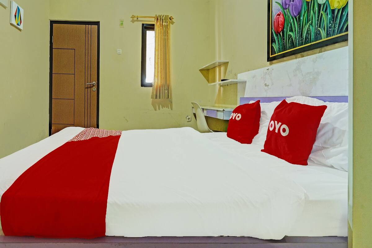 Una cama con colcha roja y blanca, dos almohadas rojas con el texto 'YOYO', una ventana con cortina, una puerta y un cuadro de flores en la pared.
