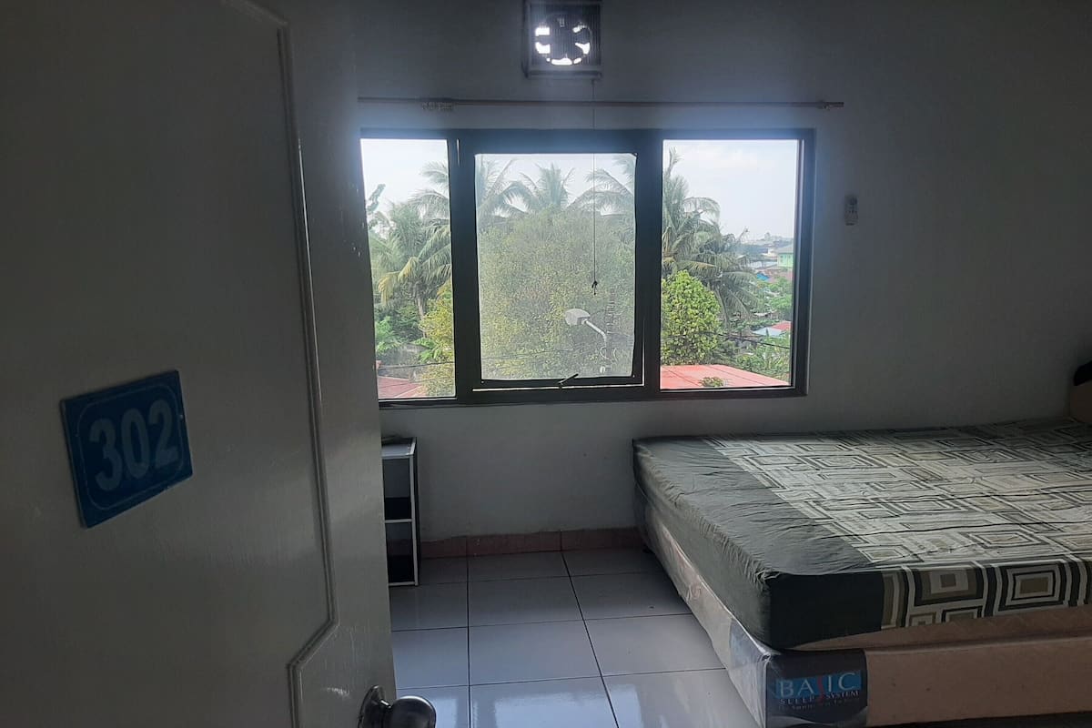 Una habitación con una cama, un ventanal con vistas a palmeras, una puerta con un cartel de número y un estante pequeño.