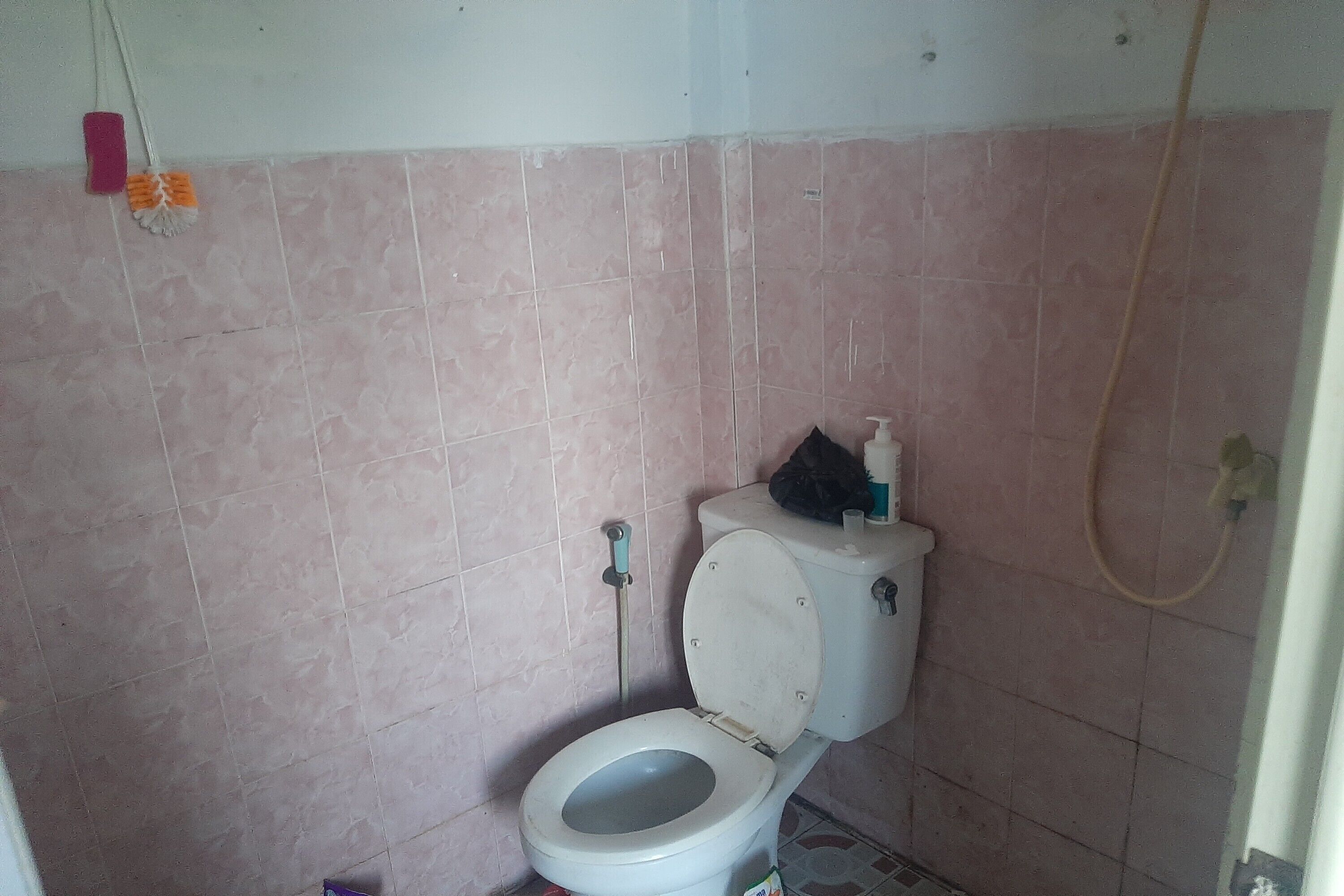 A small bathroom with pink tiles, a toilet, a shower hose, and a mop.