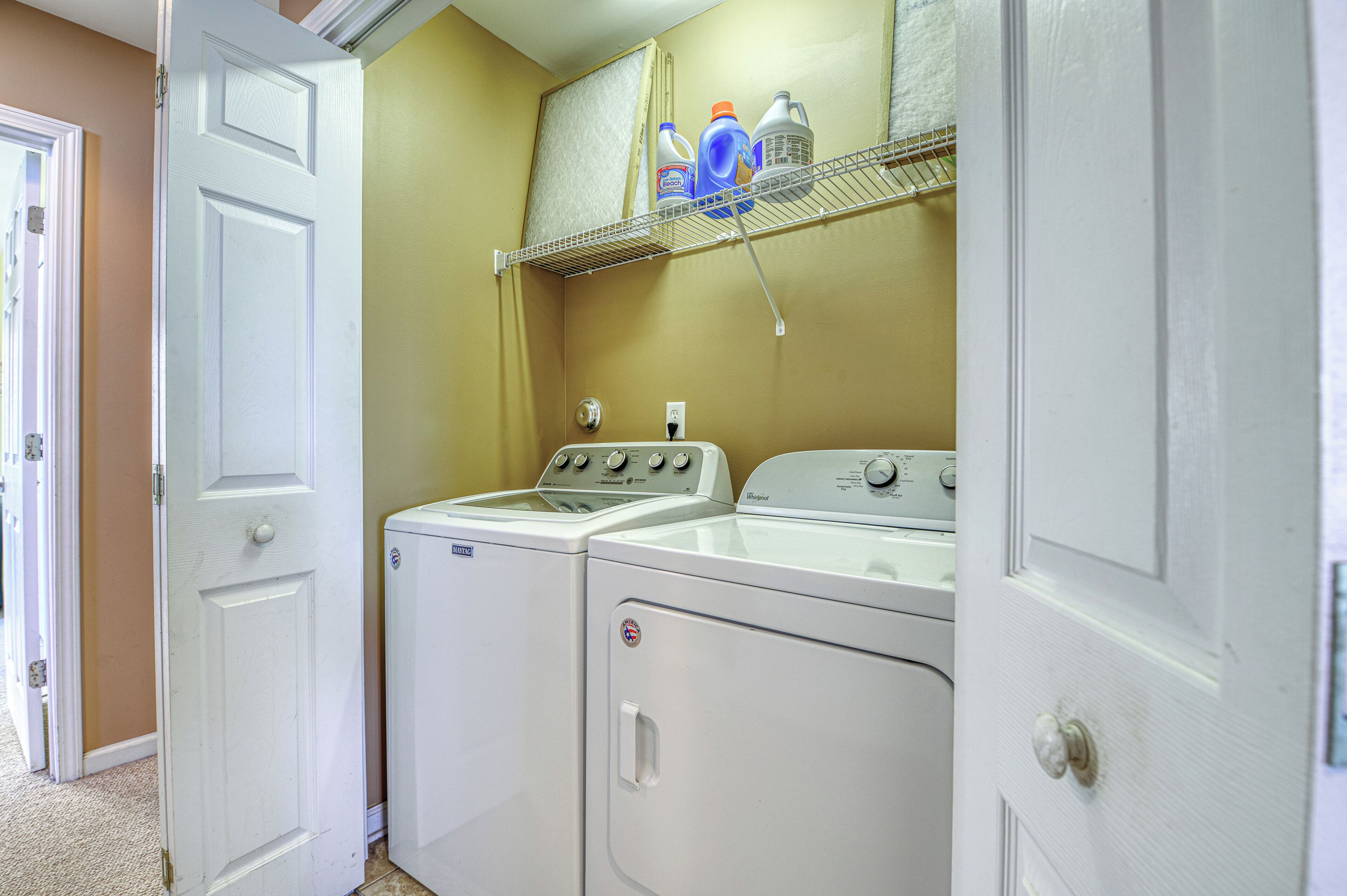A laundry room with two washers and dryers, a shelf with cleaning supplies, and a door leading to another room.