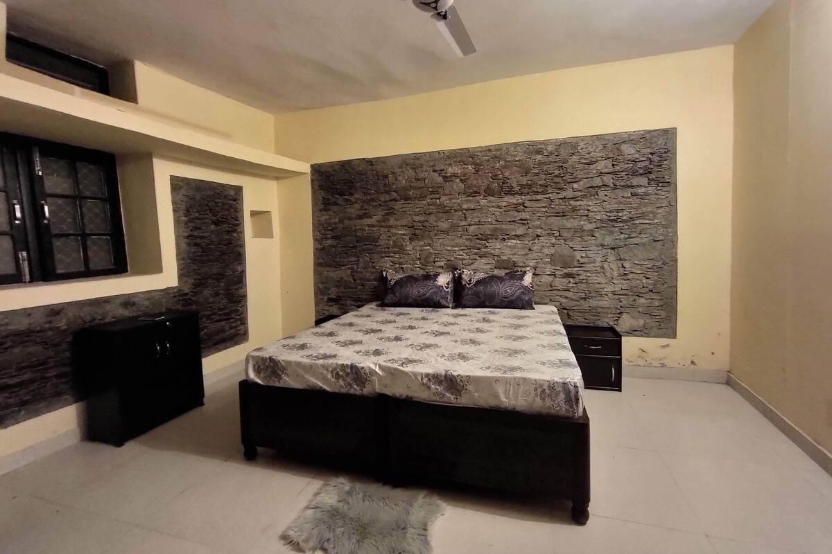 A bedroom with a stone wall, a bed with patterned bedding, a ceiling fan, and a small rug.