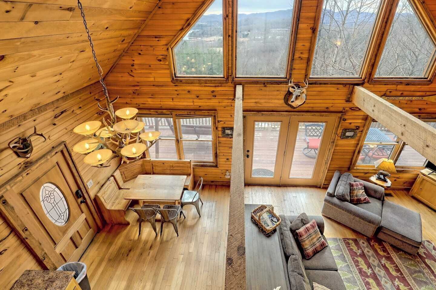 Interior de una cabaña con comedor, sofá y ventanas amplias que ofrecen vistas al exterior.