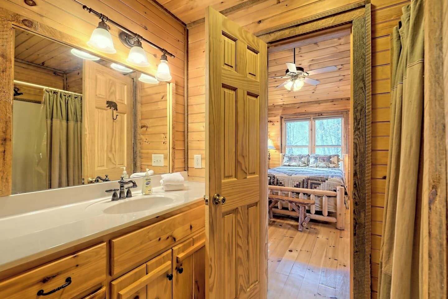 L’intérieur d’un chalet en bois comprend une salle de bain avec un grand miroir, un lavabo et un meuble-lavabo. Une chambre à coucher avec un lit et une fenêtre est visible à travers une porte ouverte.