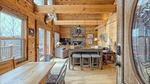 Interior acolhedor de uma cabana com área de jantar de madeira, cozinha central com lareira de pedra e uma grande janela com vista para uma paisagem nevada.