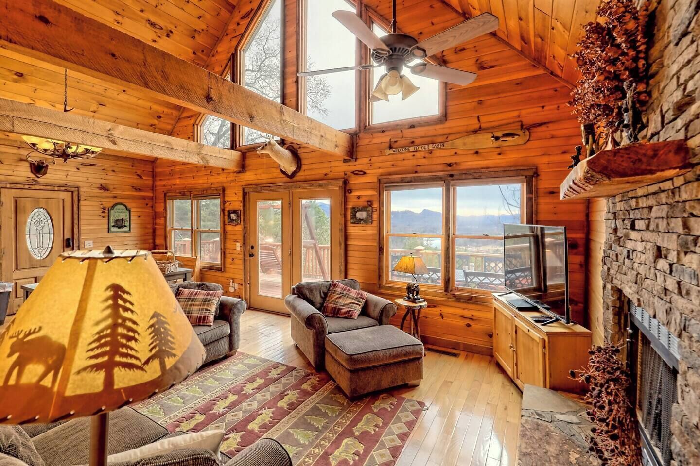 A cozy cabin interior with wooden walls, a ceiling fan, a fireplace, and a television.
