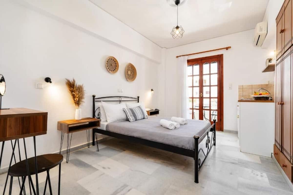 A bedroom with a bed, bedside table, chair, and a small desk. There is a window with curtains and a door leading to the bathroom.